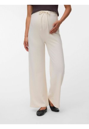 Femme portant un pantalon crème à jambes larges en tissu texturé avec une taille à cordon, associé à des ballerines noires et un haut foncé.
