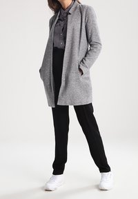 Cardigan lungo grigio con finitura testurizzata, indossato sopra una camicia button-up scura e pantaloni neri, abbinato a sneaker bianche.
