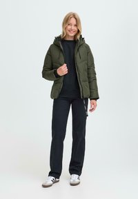 Grön vadderad jacka med huva, med dragkedjestängning och sidofickor, bärs över en svart topp och matchas med svarta byxor och sneakers.