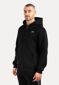 Sort zip-up hoodie lavet af blødt materiale, med en frontlomme og hætte med snoretræk. Inkluderer et lille hvidt logo på brystet.