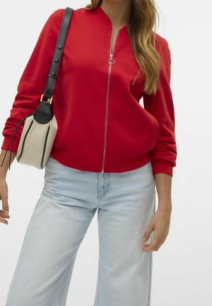 Femme portant une veste rouge zippée, un jean bleu clair et un sac à épaule beige et noir sur fond blanc.