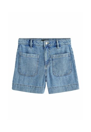 Femme portant un top sans manches en denim et un short en denim, debout en plein air près d'un bâtiment et d'arbres par une journée ensoleillée.