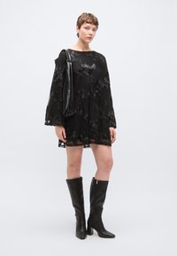 Μαύρο φόρεμα με μακριά μανίκια, floral κέντημα και άνετη εφαρμογή. Συνδυάζεται με μαύρες μπότες μέχρι το γόνατο. Κρατάει μια μαύρη τσάντα με υφή.