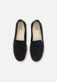 Juodos suede slip-on bateliai su apvalia nosimi, turintys jūros žole grindinį ir šviesiai beige vidinę pamušalą. Prekės ženklas "TOMS" matomas viduje.