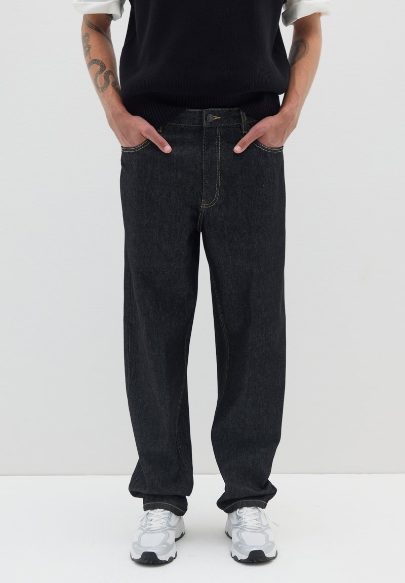 Pantalones de mezclilla negros con un corte relajado, costuras prominentes, bolsillos laterales y cierre de botón, combinados con zapatillas blancas.