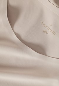 Tissu beige avec un petit logo doré et une étiquette de taille "S/M" imprimée à l’intérieur du vêtement près de l’encolure.