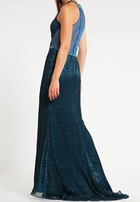 Dopasowana wieczorowa suknia w głębokim zielonym odcieniu z błyszczącym, teksturowanym materiałem. Posiada przezroczysty tył oraz ozdobny dekolt dla dodatkowego efektu.