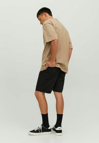 Camicia beige a maniche corte, pantaloni corti in denim neri, sneakers nere con suole bianche e calze nere. Il modello sta in piedi con le spalle rivolte, mani in tasca.