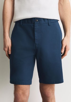 Mand iført marineblå skræddersyede shorts og en hvid ærmeløs skjorte, stående med hænder afslappet langs siderne.