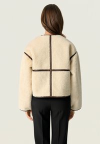 Donna che indossa un giubbotto di shearling color crema con dettagli in cucitura marrone scuro e pantaloni neri, in piedi di spalle contro uno sfondo neutro.