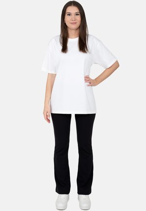 Młoda kobieta o długich brązowych włosach, ubrana w prostą białą, luźną koszulkę, czarne spodnie i białe trampki, stoi z ręką na biodrze.