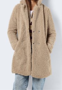 Cappotto beige in pile soffice con cappuccio, caratterizzato da chiusura con bottoni e due tasche laterali, abbinato a un maglione a righe sotto.