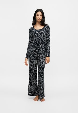Kvinde står barfodet iført et sort langærmet pyjamasæt med små hvide blomstermønstre på en ensfarvet baggrund.