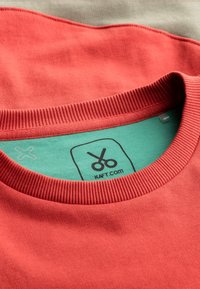 Rode katoenen T-shirt met geribbelde halslijn, met een groene label aan de binnenkant bedrukt met scharen en "kaft.com." Twee shirts op de achtergrond.