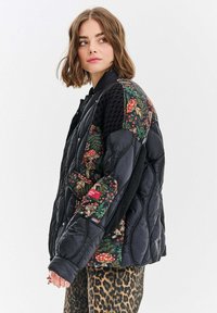 Czarna pikowana kurtka z rękawami w kwiatowy wzór, o siatkowej teksturze i czerwonych akcentach. Nosi na sobie spodnie w panterkę.