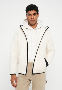 Valkoinen fleece-takki mustilla yksityiskohdilla, vetoketju suljettuna ja sivutaskuilla. Päällä vaalea huppari ja yhdistettynä vaaleisiin housuihin. Sileä pinta.