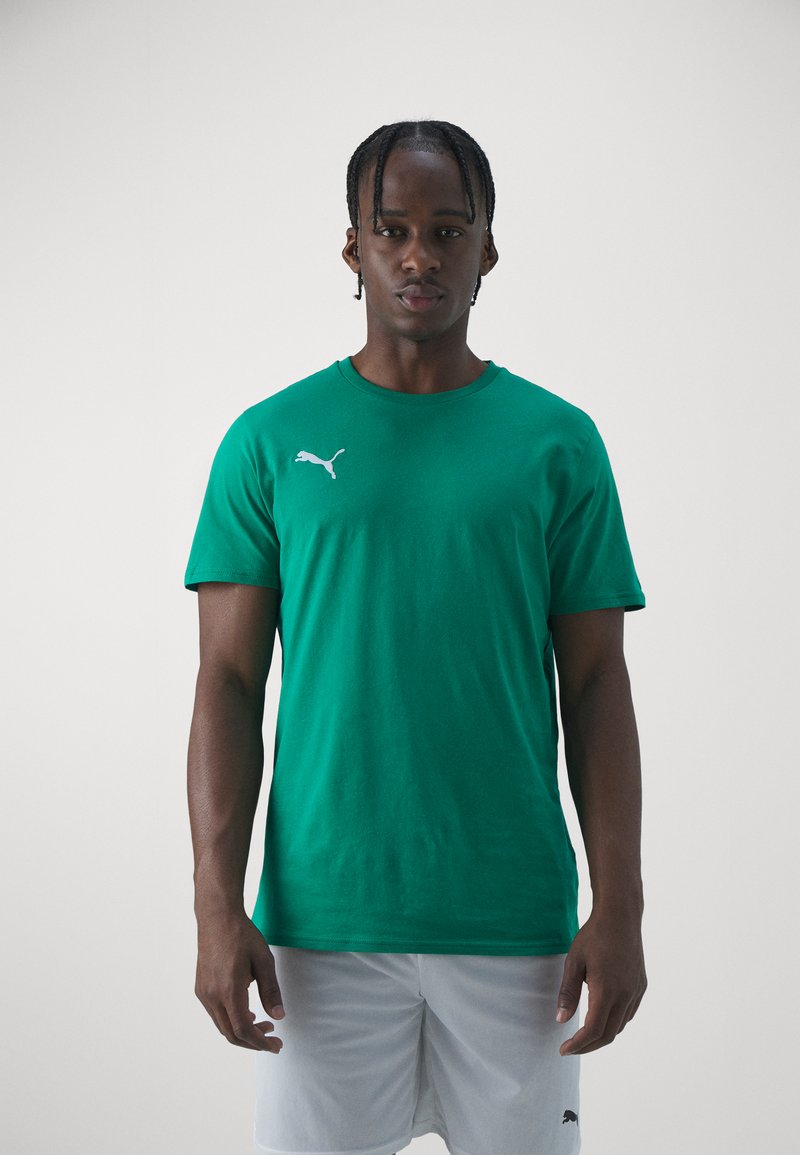 Zeleno bombažno majico s kratkimi rokavi krasi belo Puma logo na levem prsnem delu. Model nosi bele kratke hlače.