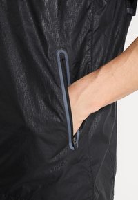 Main insérée dans une poche zippée sur une veste noire Nike présentant un motif en relief du texte "Just Do It" et un contour de fermeture éclair réfléchissant.