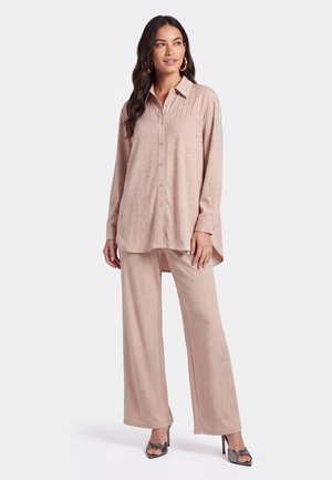 Donna che indossa una camicia lunga beige con bottoni e pantaloni ampi abbinati con piccoli motivi a pois, abbinati a tacchi aperti scuri.