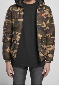 Kurtka puchowa w kamuflażu w zielonych, brązowych i czarnych odcieniach, z zamkiem z przodu, elastycznymi mankietami i pomarańczową naszywką z logo na piersi.