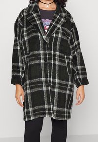 Zwart en groene geruite oversized jas met een geknipte kraag, twee voorzakken en knoopsluiting. Stof lijkt zacht en heeft een gestructureerde uitstraling.