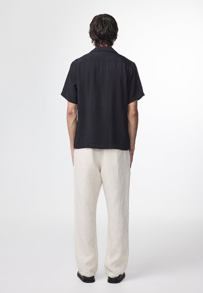 Svart kortärmad skjorta i texturerad tyg, kombinerad med lösa, ljusa beige linbyxor. Svarta skor fulländar outfiten.