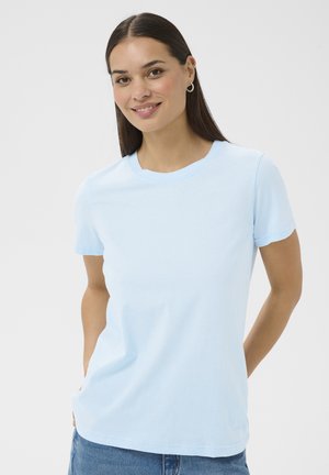 Smilende kvinde med glat mørkt hår iført lyseblå kortærmet t-shirt og øreringe, stående foran en ensfarvet hvid baggrund.