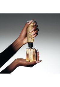 Klar glasflaska för parfym i handen, fylld med gyllene vätska. Svart bakgrund framhäver dess geometriska, moderna design och detaljerade etikett.