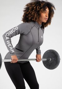 Grijze langekater mouw trainingsshirt met een ritssluiting aan de nek en een zijzak, met witte tekst langs de mouw. Zwarte legging en gewichten zijn zichtbaar.