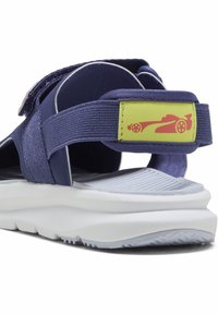Mørkeblå sandal med justerbare stropper, udstyret med et fremtrædende gult mærke med et rødt racerbilsdesign og en struktureret hvid sål.