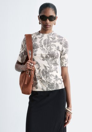 Femme portant un haut à manches courtes imprimé floral, jupe noire, lunettes de soleil foncées, boucles d'oreilles créoles, portant un sac à bandoulière en daim marron.