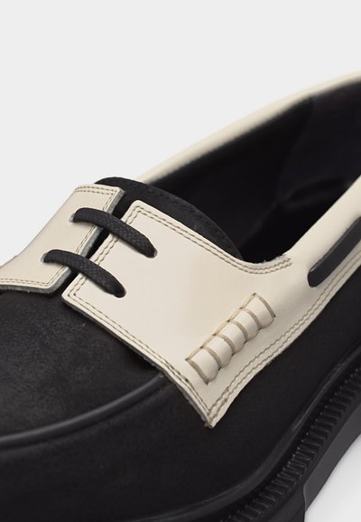 Μονόχρωμο μαύρο και άσπρο μοκασίνι με μαύρα κορδόνια περασμένα μέσα από λευκή δερμάτινη λωρίδα ραμμένη πάνω στο μαύρο πάνω μέρος του παπουτσιού.