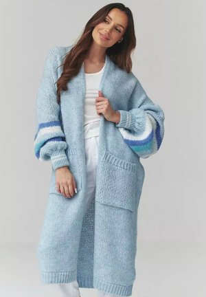 Kobieta ubrana w długi, jasnoniebieski, dziergany kardigan z dużymi kieszeniami i wielokolorowymi paskowanymi rękawami, na białej bluzce i spodniach.