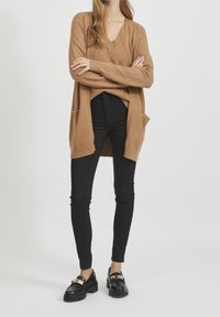 Cárdigan oversized color camello con cuello en V, dos bolsillos delanteros, llevado sobre una camiseta negra y jeans ajustados negros, combinado con mocasines negros.