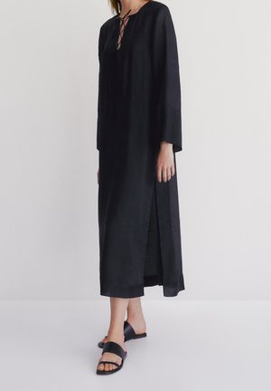 Femme portant une longue robe noire avec un décolleté à lacets, une fente latérale et des manches longues, assortie de sandales plates noires.