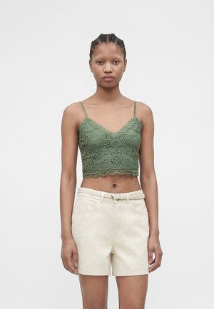 Donna che indossa una canottiera corta in pizzo verde con spalline sottili e pantaloncini crema a vita alta con cintura intrecciata, in piedi davanti a uno sfondo semplice.