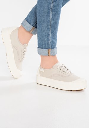 Person iført lyse beige platform-sneakers med hvide såler og opsmøgede blå jeans, stående på et hvidt gulv.