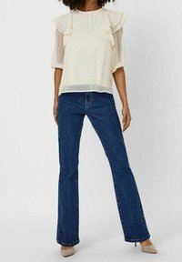 Cremefarvet bluse med gennemsigtige ærmer, flæse detaljer og prikker; parret med mørkeblå flagrende jeans og beige hæle.