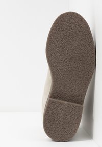 Zapato de ante beige con suela de goma texturizada, con punta redonda y tacón plano, apoyado sobre un fondo blanco.