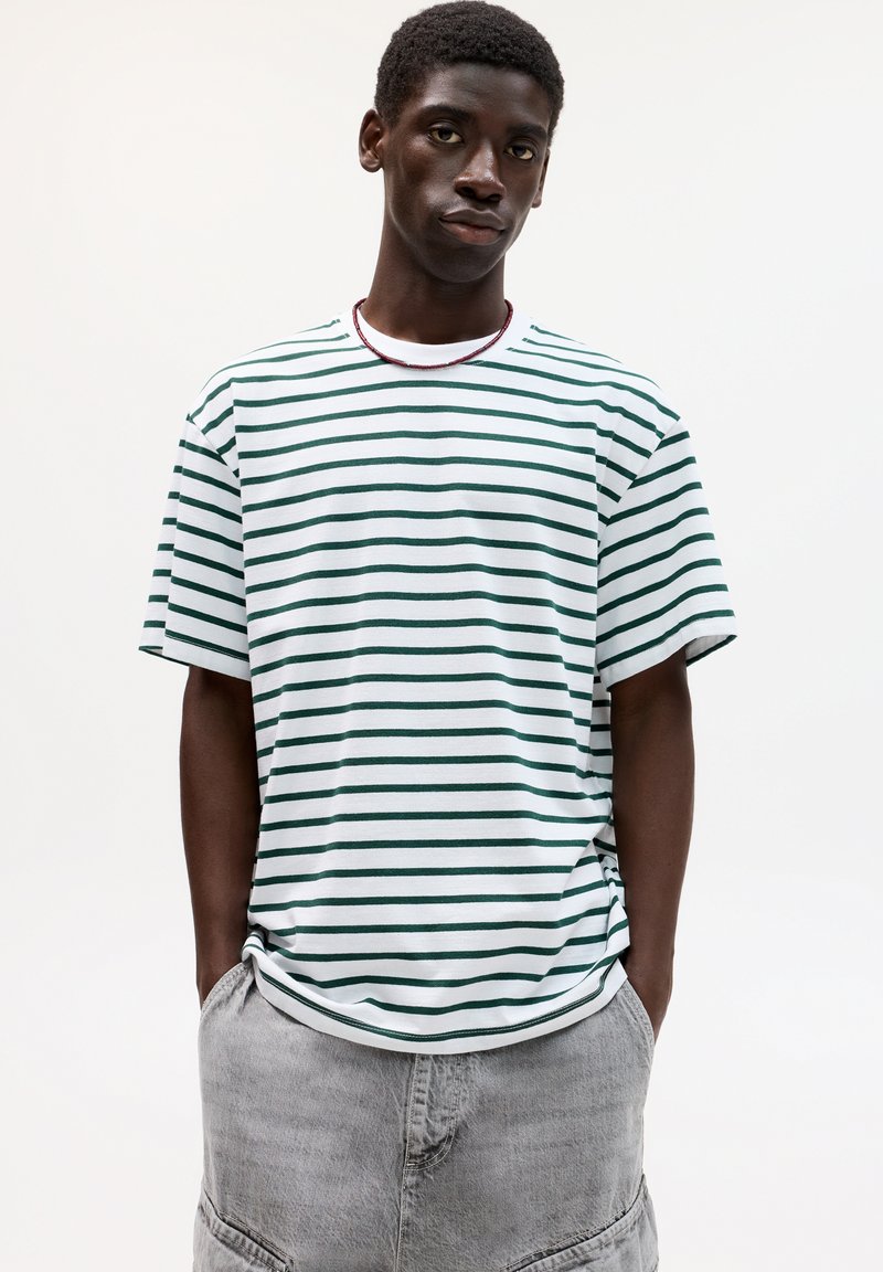 Tricou alb cu dungi orizontale verzi închise, guler rotund cu tiv contrastant, mâneci scurte, croială lejeră, purtat cu pantaloni scurți gri.