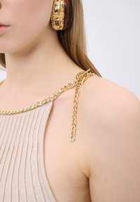 Beige geribbelde stof top met een gouden kettingdetail op de schouder, vergezeld van een textuur gouden oorbel.