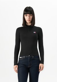 Vrouw met steil, donker haar in een aansluitend zwart longsleeve shirt met een klein logo en donkerblauwe jeans tegen een witte achtergrond.