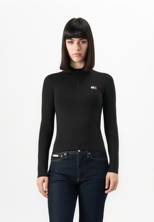 Kvinne med rett, mørkt hår iført en tettsittende svart langermet topp med en liten logo og mørkeblå jeans foran en hvit bakgrunn.