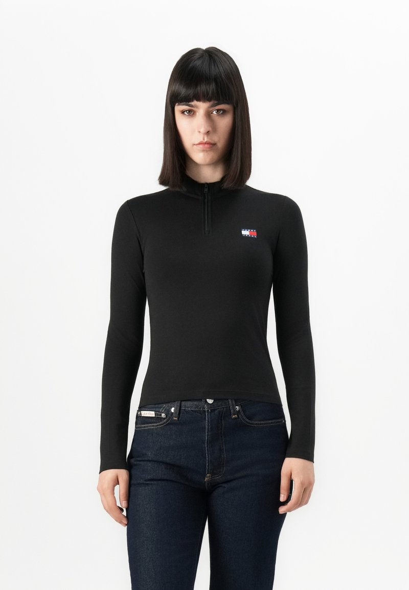 Vrouw met steil, donker haar in een aansluitend zwart longsleeve shirt met een klein logo en donkerblauwe jeans tegen een witte achtergrond.