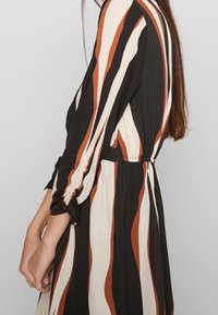 Gestreiftes Kleid in Schwarz, Weiß und Rostfarben; mit tailliertem Schnitt, langen Ärmeln und gerafften Details an den Bündchen.