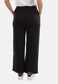 Persona che indossa pantaloni neri a gamba larga con cintura elastica e sneaker bianche con dettagli neri sul tallone, in piedi con le mani nelle tasche.