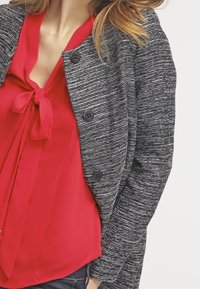 Svart- och vitrandig cardigan med tre stora knappar, lager-på-lager över en röd blus med rosett vid kragen.