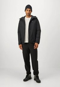 Black puffer jacket with a hood, zip closure, and side pockets over a white shirt, paired with black pants and a dark beanie.