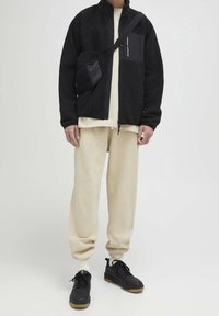 Černá fleecová bunda se zipem, béžová košile pod ní, odpovídající béžové tepláky, černé tenisky s gumovou podrážkou a malá černá crossbody taška.