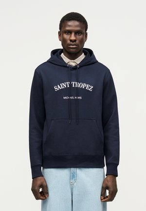 Tmavokožý muž oblečený v námornícky modrej mikine s nápisom „Saint Tropez Michael Kors“, svetlá košeľa pod ňou a svetlomodré džínsy, stojaci pred bielym pozadím.